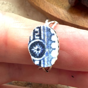 May include: A ring featuring an oval-shaped piece of white and blue patterned ceramic shard set in a silver-toned metal band. The ceramic fragment displays a dark blue circular floral motif on the left and a scalloped edge pattern on the right.