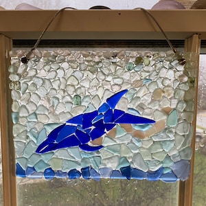 May include: A rectangular mosaic art piece featuring a blue dolphin leaping through ocean waves. The dolphin is crafted from vibrant blue glass pieces, set against a backdrop of white, light blue, and green glass fragments. A brown cord is attached for hanging.