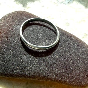May include: A simple, thin, polished silver-coloured metal ring with a smooth, rounded band. The ring features a continuous circular shape with a reflective, metallic surface that catches the light, showcasing a clean and understated design.
