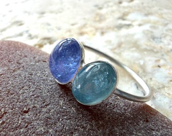 May include: A silver ring featuring two oval-shaped cabochon gemstones set side-by-side. One stone is a deep violet-blue, while the other is a lighter, translucent teal-blue. The band is a simple, smooth silver wire that curves around the finger.