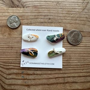 May include: Four colorful ceramic buttons with white backgrounds and a hole in the center. The buttons are arranged on a white card with the text "Collected where ever Randi travels #5434 oonamadethis.etsy.com".