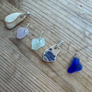 May include: Five pendants crafted from sea glass and pottery, each with a silver-tone metal loop. The pendants are in diverse shapes and colours, including a light beige stone, lavender, aqua, blue and white patterned pottery, and a deep blue sea glass.