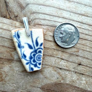 May include: A handmade pendant featuring a fragment of ceramic with a blue floral design on a cream background. The pendant has a silver bail. A coin is next to the pendant for size comparison. The pendant is on a wooden surface.