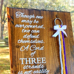 May include: A rectangular wooden board with a natural grain finish featuring white text that reads: Though one may be overpowered, two can defend themselves. A Cord of THREE strands is not easily broken. A silver ring with a white bow and three hanging cords.