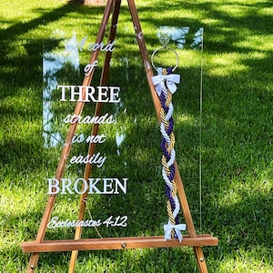 May include: A clear acrylic sign with the text "A cord of three strands is not easily broken, Ecclesiastes 4:12". A white braided rope with gold and purple strands is hanging from the sign.