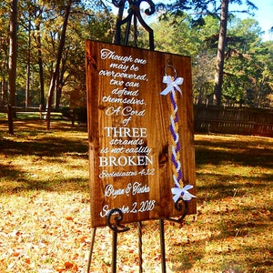 May include: A rectangular wooden sign with white text reading "Though one may be overpowered, two can defend themselves. A Cord of THREE strands is not easily BROKEN Ecclesiastes 4:12 Bryan & Tiesha September 2, 2018" and a braided cord with white, gold, and purple strands.