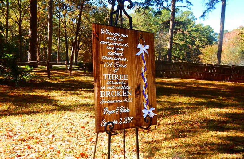 May include: A rectangular wooden sign with white text reading "Though one may be overpowered, two can defend themselves. A Cord of THREE strands is not easily BROKEN Ecclesiastes 4:12 Bryan & Tiesha September 2, 2018" and a braided cord with white, gold, and purple strands.