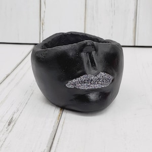 May include: A black, round, decorative planter with a face design. The planter features a pair of lips with silver glitter. The planter is made of a matte black material and sits on a white wooden surface.