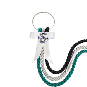 May include: A silver metal ring attached to a white satin ribbon bow featuring a small blue and silver anchor charm. Three twisted ropes in teal, silver, and black hang from the ring, creating a decorative nautical accessory with a clean design.