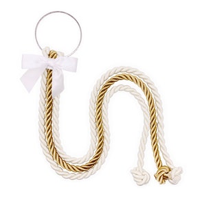 May include: A decorative hanging ornament featuring a silver-toned metal ring attached to three twisted ropes in alternating cream and metallic gold colors. A white satin ribbon is tied into a bow at the top, and the rope ends are finished with knots.