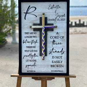 May include: A white wooden sign with a black frame and a black cross with a purple, white, and gold cord wrapped around it. The sign has the text "Estrella & Joseph" and "October 15, 2025" at the top. The sign also has the text "They are no longer two, but one flesh. Therefore what God has joined together, let no one separate." and "Matthew 19:6" at the bottom left. The sign also has the text "A cord of three strands is not easily broken" and "Ecclesiastes 4:12" at the bottom right.