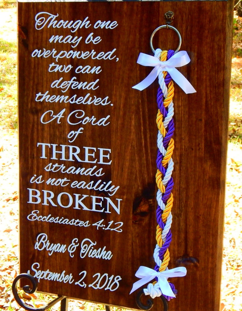May include: A dark wood rectangular plaque featuring white text that reads: Though one may be overpowered, two can defend themselves. A Cord of THREE strands is not easily BROKEN Ecclesiastes 4:12 Bryan & Tiesha September 2, 2018. A braided cord hangs from a metal ring.