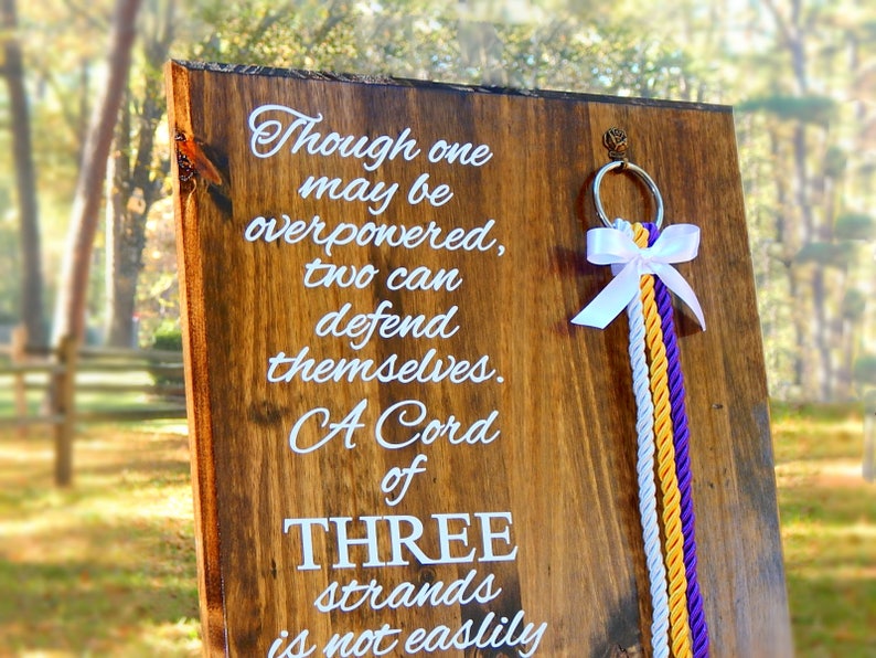 May include: A rectangular wooden board with a natural grain finish featuring white text that reads: Though one may be overpowered, two can defend themselves. A Cord of THREE strands is not easily broken. A silver ring with a white bow and three hanging cords.