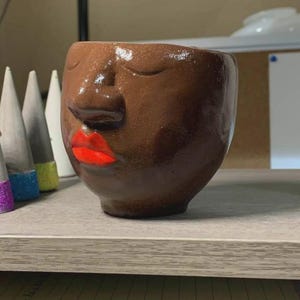 May include: A brown ceramic face planter with closed eyes and bright red lips. The handmade pot has a rounded shape and is sitting on a shelf. The planter is a unique piece of home decor.