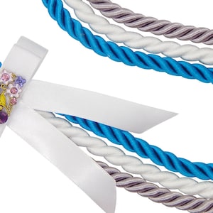 May include: Close-up of decorative rope with a white satin ribbon and a floral embellishment. The rope is in shades of blue, white, and gray. The embellishment features pink, purple, and gold accents.