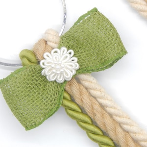 May include: A decorative bow featuring a green burlap bow, a white floral embellishment, and a collection of braided ropes in cream, tan, and green. The bow is attached to a silver ring.