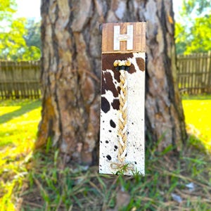 May include: A brown wooden sign with the letter "H" in white on it. The sign is attached to a piece of cowhide with a braided rope hanging from it.