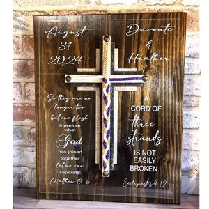 May include: A wooden plaque with a clear acrylic overlay featuring a cross with a braided cord in purple, gold, and white. The plaque has the text "August 31, 2024 Davonte & Heather" at the top, and the text "So they are no longer two but one flesh therefore what God has joined together let no one separate. Matthew 19 6" on the left side. The right side has the text "A CORD OF three strands IS NOT EASILY BROKEN. Ecclesiastes 4 12".