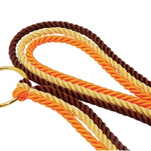 May include: A decorative cord featuring three twisted strands in dark brown, light cream, and bright orange, all secured to a polished gold-tone metal ring. The braided ropes are arranged in loops, showcasing a smooth, shiny texture throughout.