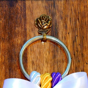 May include: A silver-toned metal ring attached to a bronze-colored rose ornament. The ring is adorned with three small, twisted fabric ribbons in white, yellow, and purple, positioned at the bottom, with a larger white ribbon draped below.