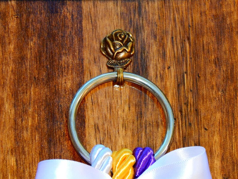May include: A silver-toned metal ring attached to a bronze-colored rose ornament. The ring is adorned with three small, twisted fabric ribbons in white, yellow, and purple, positioned at the bottom, with a larger white ribbon draped below.