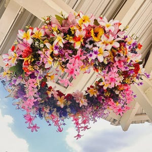 May include: A vibrant floral wreath composed of a variety of colorful flowers, including pink, yellow, and purple blooms. The wreath is suspended from a white structure, with a blue sky visible in the background. The flowers are arranged in a circular shape.