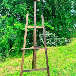 May include: A wooden easel standing on a grassy lawn. The easel is made of dark brown wood and has a simple design.