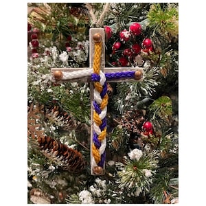 May include: A wooden cross ornament with a braided rope design in purple, gold, and white. The ornament is hanging on a Christmas tree.