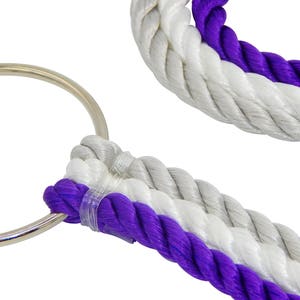 May include: A silver metal ring attached to three strands of twisted rope in alternating purple and white colors. The ropes are secured together with a clear plastic fastener near the ring, creating a durable and textured loop design.