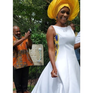 May include: A sleeveless white A-line dress featuring a side panel with yellow, blue, and gold patterned fabric. The dress is paired with a large, structured yellow headwrap. A wooden sign in the background displays the text A Cord of THREE strands.