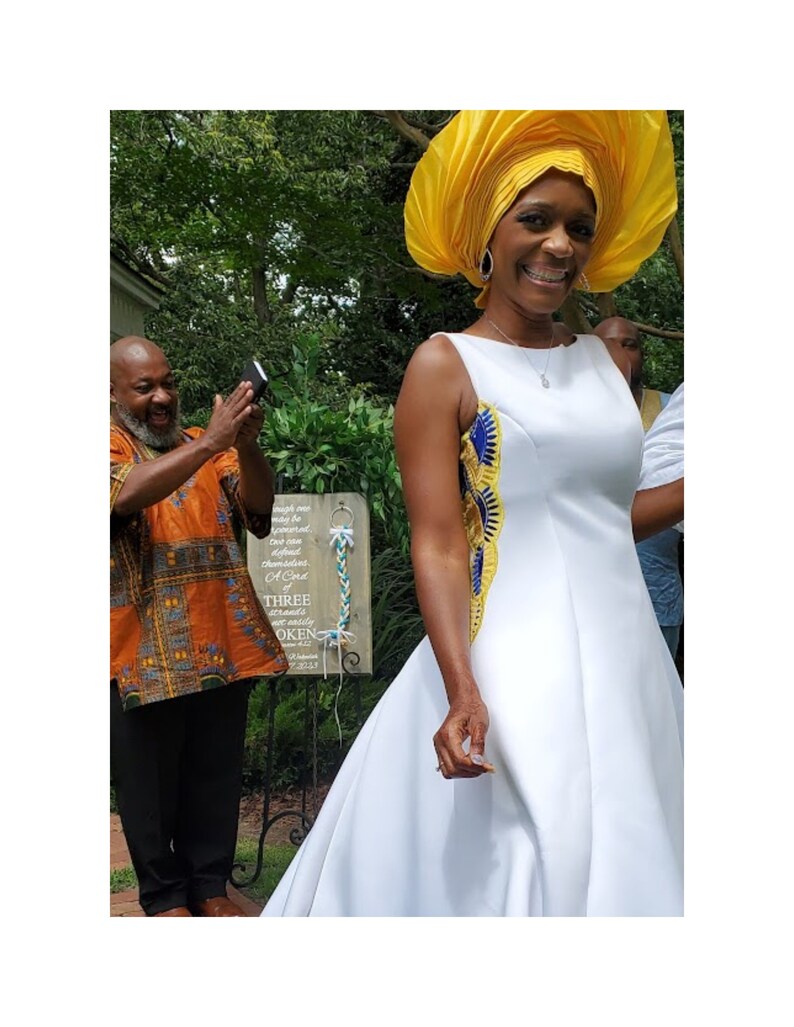 May include: A sleeveless white A-line dress featuring a side panel with yellow, blue, and gold patterned fabric. The dress is paired with a large, structured yellow headwrap. A wooden sign in the background displays the text A Cord of THREE strands.