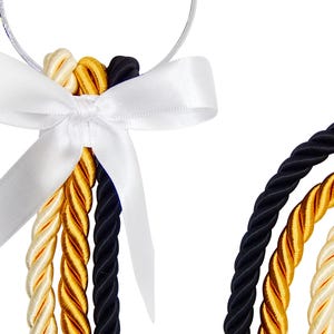 May include: Close-up of a decorative ornament featuring twisted cords in black, gold, and cream colors. A white satin bow secures the cords to a clear loop. The cords are arranged in a cascading pattern against a white background.