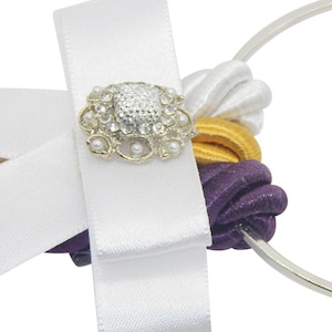 May include: A decorative wedding bouquet holder featuring a white satin ribbon with a jeweled embellishment. The holder includes white, gold, and purple accents, and a silver metal ring.
