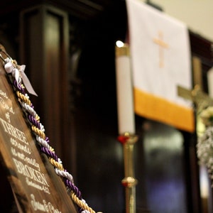 May include: A wooden sign featuring white script text that reads, Though one may be overpowered, two can defend themselves. A cord of three strands is not easily broken. Ecclesiastes 4:12. A braided cord in purple, gold, and white is attached.