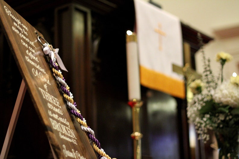 May include: A wooden sign featuring white script text that reads, Though one may be overpowered, two can defend themselves. A cord of three strands is not easily broken. Ecclesiastes 4:12. A braided cord in purple, gold, and white is attached.