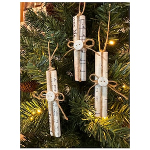May include: Three handmade Christmas ornaments. Each ornament is a rolled cylinder of vintage sheet music, tied with twine and adorned with a white button. The ornaments hang from a Christmas tree with green needles and small lights.