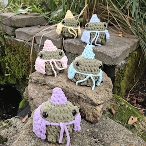 May include: Five handmade crochet frog figurines in various pastel colors, including yellow, pink, blue, and purple. The frogs have black eyes and are posed on a stone surface, surrounded by greenery. Each frog has a unique hat and scarf.