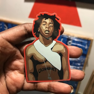 May include: A sticker of a man with dreadlocks, a white bandana, and a tattoo that says "Cash Money" on his chest. The sticker is red and has a white background.