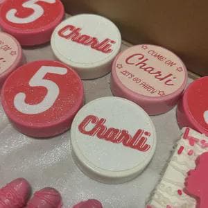 May include: Close-up of several pink and white decorative items. Some items are round with the number "5" or the name "Charlie" in red. Other items are shaped like ice cream cones and a cake, all in shades of pink.