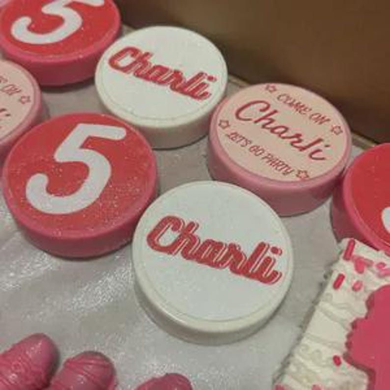 May include: Close-up of several pink and white decorative items. Some items are round with the number "5" or the name "Charlie" in red. Other items are shaped like ice cream cones and a cake, all in shades of pink.