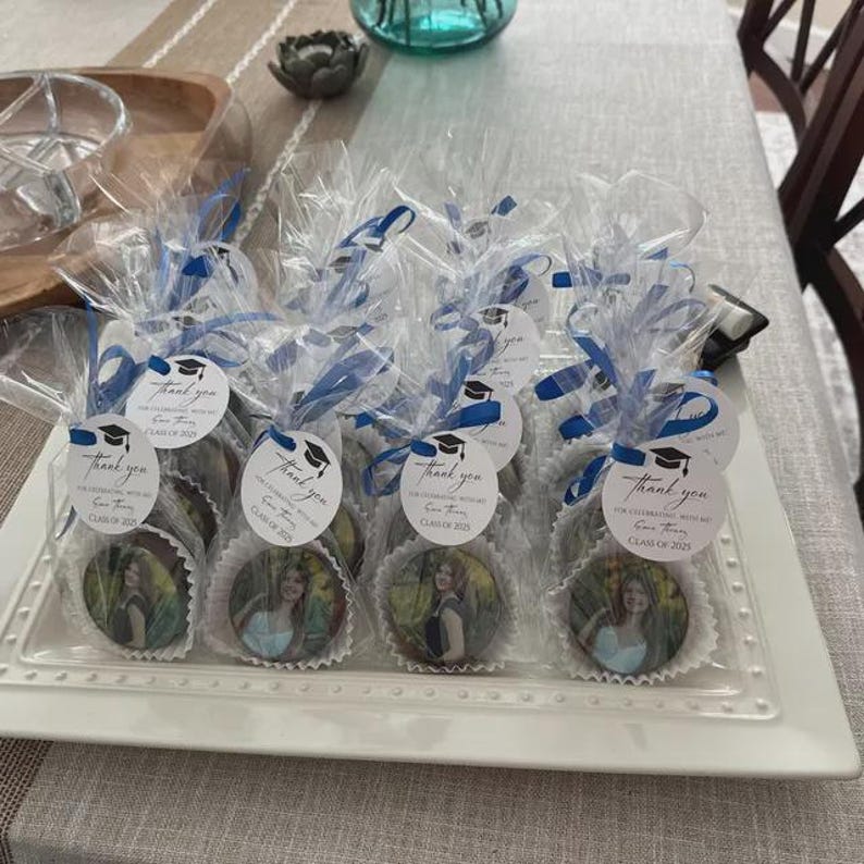 May include: A white rectangular platter holds individually wrapped treats. Each treat is adorned with a photo and a circular tag with text and a graduation cap graphic. The treats are wrapped in clear cellophane and tied with blue ribbon.