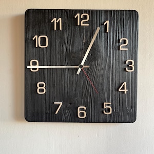 Könnte beinhalten: Eine schwarze Holzuhr mit weißen römischen Ziffern und silbernen Zeigern. Das Zifferblatt der Uhr ist aus recyceltem Holz mit rustikalem Finish.