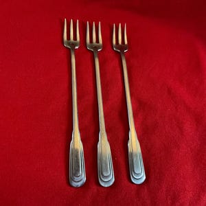 May include: Three silver-toned metal cocktail forks with long, slender handles and three tines. Each fork features a stepped, rectangular design at the base of the handle. The forks are arranged vertically in a parallel row, showcasing a metallic finish.