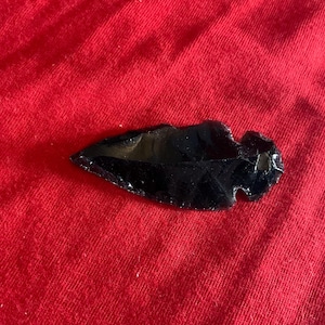 May include: A black obsidian arrowhead with a sharp, pointed tip and a notched base. The surface of the stone is smooth and reflective, showing a glossy, dark appearance with subtle flaked edges along the sides of the pointed projectile point.