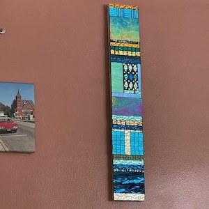 Puede incluir: Una pieza de arte de pared de mosaico vertical con un esquema de color azul, verde y dorado. El mosaico está hecho de pequeñas baldosas cuadradas dispuestas en un patrón geométrico.