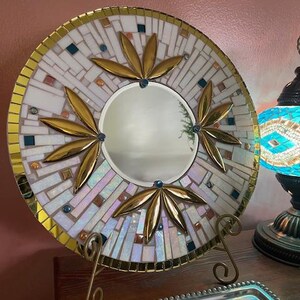 May include: A round mirror with a white and gold mosaic design. The mirror is surrounded by a gold frame and has three gold leaf designs around the centre. The mirror is on a gold stand.