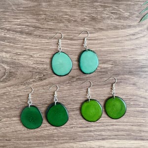 May include: A set of six earrings featuring green and teal wood slices with silver hooks. The earrings are arranged in two rows of three.