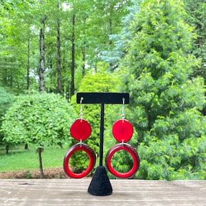 May include: A pair of red wooden earrings with a circle design. The earrings are hanging from a black earring stand.