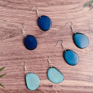 May include: Five pairs of earrings with oval-shaped blue and teal resin stones. The earrings are on a wooden surface.