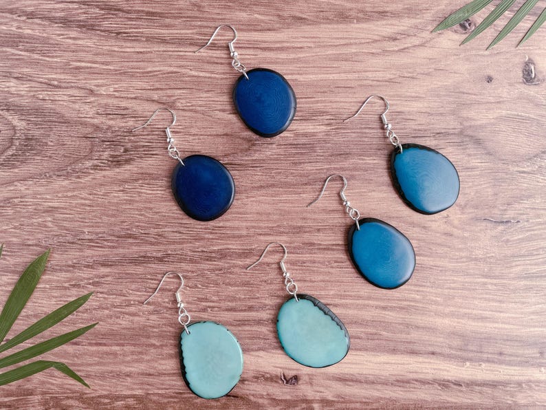 May include: Five pairs of earrings with oval-shaped blue and teal resin stones. The earrings are on a wooden surface.